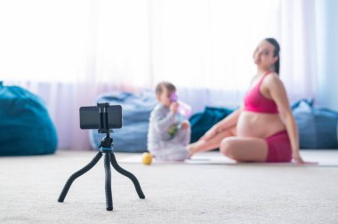 Hamile bir kadın oğluyla jimnastik yapıyor. Müstakbel anne telefonda blogu için bir eğitim videosu çekiyor. Bebekli fitness.