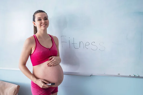 Těhotná žena ve sportovním oblečení píše slovo fitness na bílou tabuli. Holka ve třetím trimestru chodí na sport s holým bříškem. Kurzy pro nastávající matky. — Stock fotografie