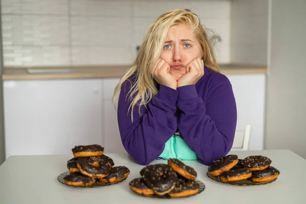 Sorglig fet blondin tittar på munkarna med chokladglasyr. En överviktig ung kvinna sitter i köket och tvivlar på sitt val. Flicka på diet. — Stockfoto