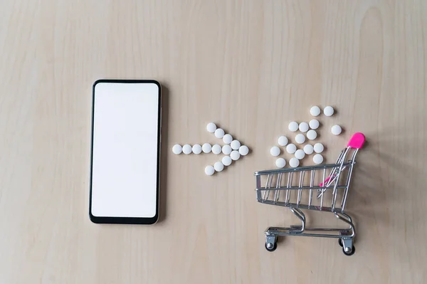 Online pharmacy concept. Buying medicine online over the phone. Delivery of pharmaceuticals. Mini trolley with tablets and smartphone with white blank screen.