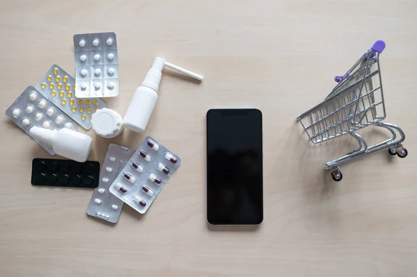 Online έννοια φαρμακείο. Αγοράζοντας φάρμακα σε απευθείας σύνδεση από το τηλέφωνο. Παράδοση φαρμακευτικών προϊόντων. Μίνι τρόλεϊ με διαφορετικά δισκία, σπρέι, κάψουλες και ένα smartphone με μαύρη λευκή οθόνη. — Φωτογραφία Αρχείου