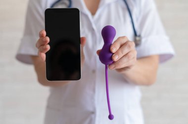 Meçhul jinekolog kadın sağlığını korumak için kegel simülatörü öneriyor. Doktor telefonla senkronize edilmiş bir vibratör gösteriyor. Pelvik zemin kasları için bir cihaz..