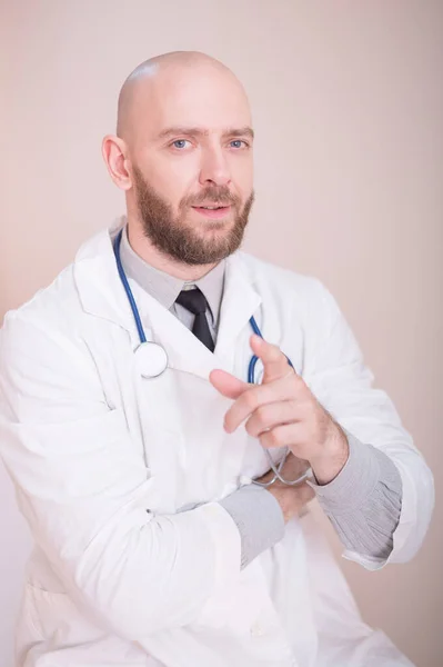 Un médico masculino se sienta en un escritorio en un consultorio médico. Un hombre calvo con barba trabaja como terapeuta en una clínica. Cardiólogo amigable con un estetoscopio en una bata de laboratorio. Foto vertical . — Foto de Stock