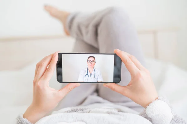 Μια απρόσωπη γυναίκα ξαπλώνει ανάσκελα στο κρεβάτι της στο σπίτι και κοιτάζει τα τηλεφωνικά βίντεο από τον γιατρό. Ο θεράπων ιατρός δίνει online συμβουλές στον ασθενή σχετικά με τη θεραπεία του coronavirus. — Φωτογραφία Αρχείου