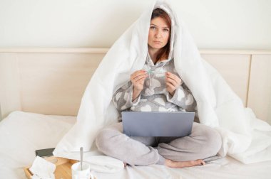 Yataktaki kadın kafasıyla battaniyeyle örtülmüş ve sıcaklığı ölçüyor. Soğuk algınlığı olan bir kız dizlerinin üzerinde dizüstü bilgisayarla yatak odasında oturuyor..