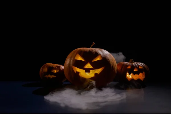 Halloweenská karta. Jack o lucerna se svíčkami zářit na černém pozadí. Řada strašidelných dýní s vyřezávanými grimasami kouří ve tmě. — Stock fotografie