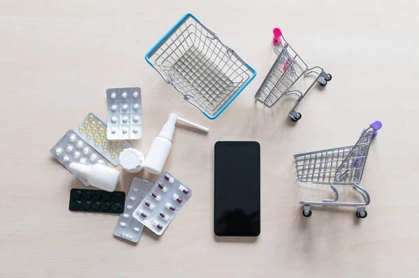 Online pharmacy concept. Buying medicine online over the phone. Delivery of pharmaceuticals. Mini trolley with different tablets, sprays, capsules and a smartphone with a black blank screen.