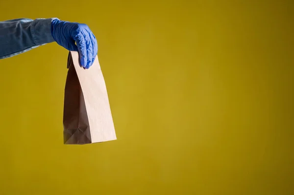 Closeup of female hands in gloves and a denim shirt. Delivery man holds an empty paper bag on a yellow background. Craft packaging for takeaway snack. Antimicrobial protection. — Stock Photo, Image