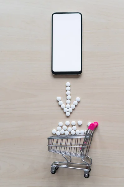 Online pharmacy concept. Buying medicine online over the phone. Delivery of pharmaceuticals. Mini trolley with tablets and smartphone with white blank screen.