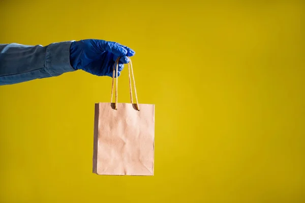 Fecho de mãos femininas em luvas e uma camisa de ganga. Um entregador está segurando um pequeno saco de papel junto às canetas em um fundo amarelo. Embalagem artesanal para lanche takeaway. Protecção antimicrobiana . — Fotografia de Stock