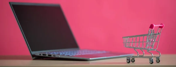 Concepto de compras online. Mini carrito de comestibles y portátil sobre un fondo rosa. Carro pequeño y ordenador personal . —  Fotos de Stock