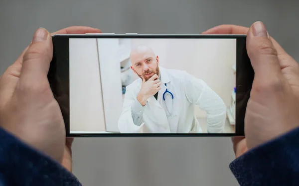 Fecho de mãos femininas com um telefone. Um médico careca consulta remotamente um paciente com uma webcam. A mulher diz com um médico amigável para chamadas de vídeo . — Fotografia de Stock