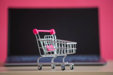 İnternetten alışveriş konsepti. Mini market arabası ve pembe arka planda dizüstü bilgisayar. Küçük araba ve kişisel bilgisayar.