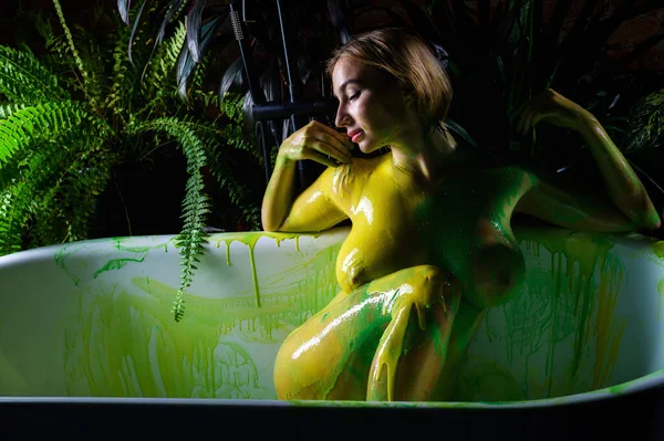 Hermosa mujer desnuda en pintura amarillo-verde sobre un fondo de plantas de interior. Una chica con pechos muy grandes toma un baño . —  Fotos de Stock