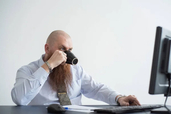 Лысый мужчина с рыжей бородой пьет кофе и работает за компьютером. Мужчина-менеджер в белой рубашке за персональным компьютером в офисе . — стоковое фото