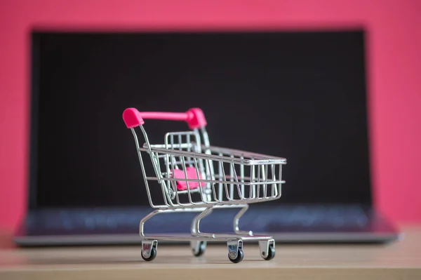 Concepto de compras online. Mini carrito de comestibles y portátil sobre un fondo rosa. Carro pequeño y ordenador personal . —  Fotos de Stock