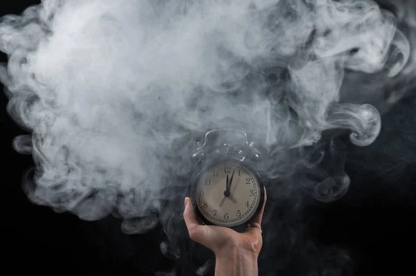 Primer plano de una mano femenina sosteniendo un reloj sobre un fondo negro en humo. Reloj despertador a medianoche en una niebla mística . —  Fotos de Stock