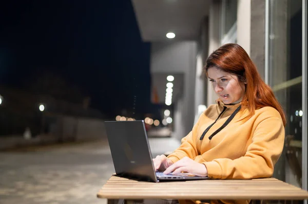 한 여성은 저녁늦게 한 여름 카페에서 노트북으로 원격으로 일을 합니다. 웃는 한 소녀가 나무 탁자에 앉아 낭비 된 거리에 앉아서 공부하고 있다. — 스톡 사진