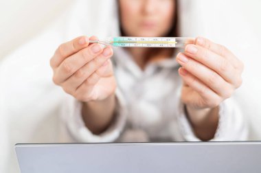 Pijamalı bir kadın kamerada cıva termometreyi gösteriyor. Ateşli yüzü olmayan bulanık kız yatakta oturuyor..