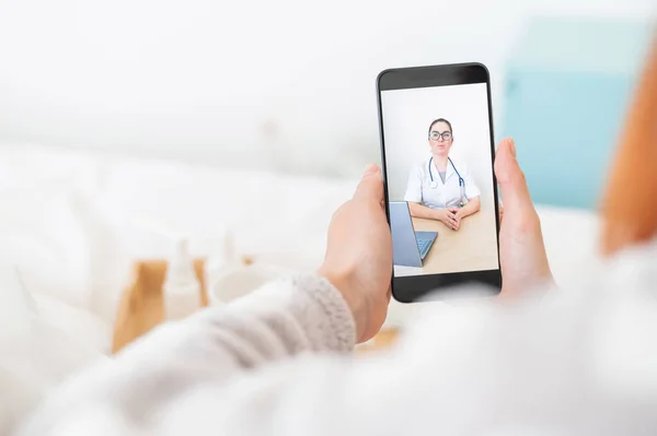 Концепція дистанційного лікування. Вид ззаду жінки в ліжку в піжамі зі смартфоном в руках. Пацієнт з грипом дивиться медичний відео блог . — стокове фото