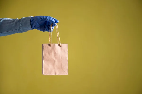 Fecho de mãos femininas em luvas e uma camisa de ganga. Um entregador está segurando um pequeno saco de papel junto às canetas em um fundo amarelo. Embalagem artesanal para lanche takeaway. Protecção antimicrobiana . — Fotografia de Stock