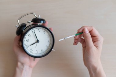 Kadının elinde hamilelik testi ve çalar saat var. Kadın doğurganlık biyolojik saati konsepti. Bir şerit.
