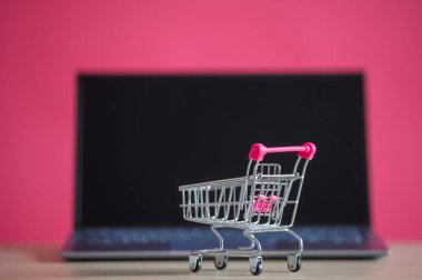 İnternetten alışveriş konsepti. Mini market arabası ve pembe arka planda dizüstü bilgisayar. Küçük araba ve kişisel bilgisayar.
