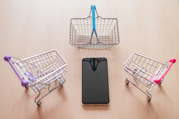 Vista superior en un teléfono móvil dos carros de comida y una cesta en una mesa de madera. Compras en línea en un smartphone. Miniaturas . —  Fotos de Stock