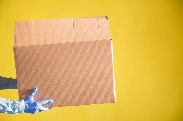 Closeup of female hands in gloves and a denim shirt. The delivery man passes a cardboard box to the customer on a yellow background. Antimicrobial protection in quarantine. Cropped. — Stock Photo, Image