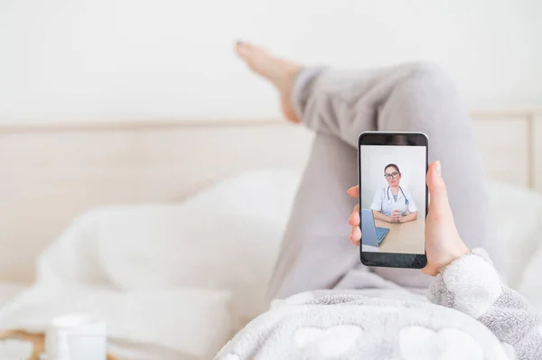 Cep telefonunun ekranında bir doktorun portresi. Çevrimiçi tıp konsepti. Evde bir kadın yatağında uzanıyor ve video bağlantısı üzerinden uzman doktorla görüşüyor.. — Stok fotoğraf