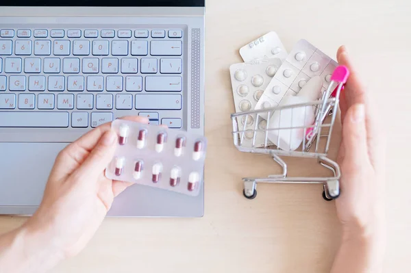 Online pharmacy. Closeup of female hands on a laptop and tablet keyboard in a mini shopping trolley with tablets. A woman chooses medicines on the Internet with home delivery.