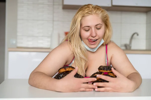 Een dikke blonde met grote borsten eet een donut met chocolade in de keuken. Een zwaarlijvige jonge vrouw heeft twee borden vol zoete gebak. Meisje met een medisch masker. — Stockfoto