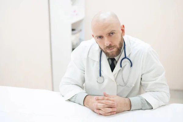 Un medico maschio si siede ad una scrivania in uno studio medico. Un uomo calvo con la barba lavora come terapista in una clinica. Cardiologo serio con uno stetoscopio in camice da laboratorio. Foto orizzontale . — Foto Stock