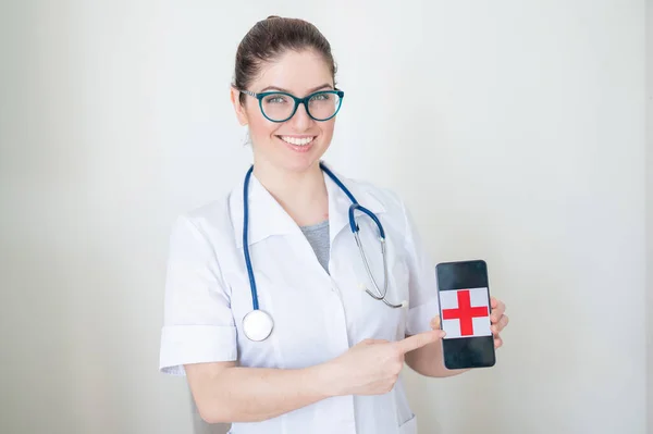 Female doctor in glasses holds a smartphone with a red cross on the screen. A nurse smiles in a medical coat with a stethoscope on her shoulders indicates a cell phone.