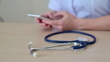 Tanımlanamayan doktor aralarda akıllı telefondan yazıyor. Cep telefonuyla online danışmanlık yapan bir kadın kardiyologun ellerinin yakın çekimi. Masaüstünde steteskop.