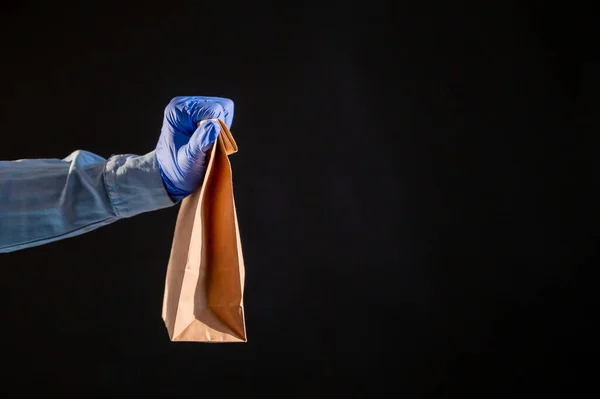 Female hand in a blue glove holds a brown paper bag on a black background. Safe food delivery to your home. A woman in a denim shirt holds out a cardboard bag to a customer. — Stock Photo, Image