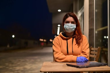 Tıbbi maskeli ve eldivenli bir kadın gece geç saatlerde yalnız yürür. Kız karantinada bir dizüstü bilgisayarla ahşap bir masada oturuyor. Coronavirüs.