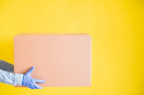 Close-up female hands in gloves holds a small cardboard box on a yellow background. Online store with home delivery. A courier in a denim shirt holds out a craft cardboard bag to a customer. — Stock Photo, Image