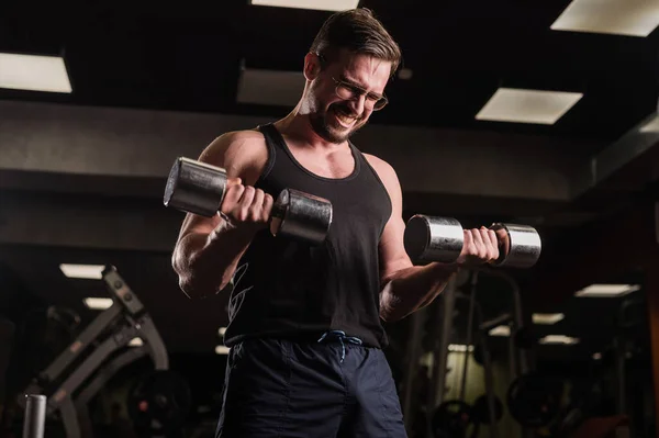 Un hombre con esfuerzo hace un ejercicio para bíceps y tríceps con una mancuerna. El culturista en relieve sonríe. Musculoso chico con gafas en el gimnasio. —  Fotos de Stock