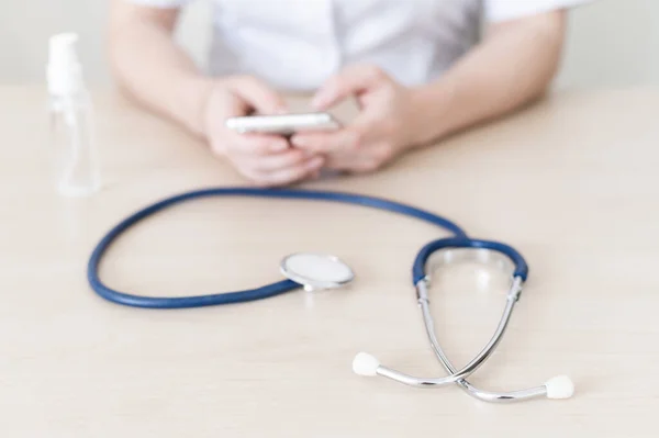 O médico está escrevendo no smartphone durante o intervalo. cardiologista feminino dá consulta on-line por telefone celular. Estetoscópio e mão anti-séptico na área de trabalho . — Fotografia de Stock