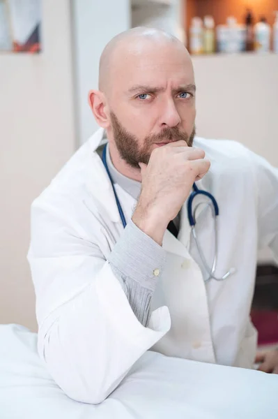 Een mannelijke dokter zit aan een bureau in een medisch kantoor. Een kale man met een baard werkt als therapeut in een kliniek. Ernstige cardioloog met een stethoscoop in een labjas. Verticale foto. — Stockfoto