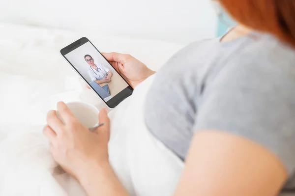 一个拿着智能手机的病假妇女。一名流感患者正在看医疗视频博客。医生通过电话进行在线会诊.卧床休息. — 图库照片