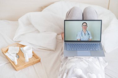 Karantina sırasında bir kadın doktoruyla iletişim kurmak için dizüstü bilgisayar kullanıyor. Kız sırt üstü uzanıyor ve tıbbi video blogu izliyor. Doktor çevrimiçi danışmanlık yapıyor..