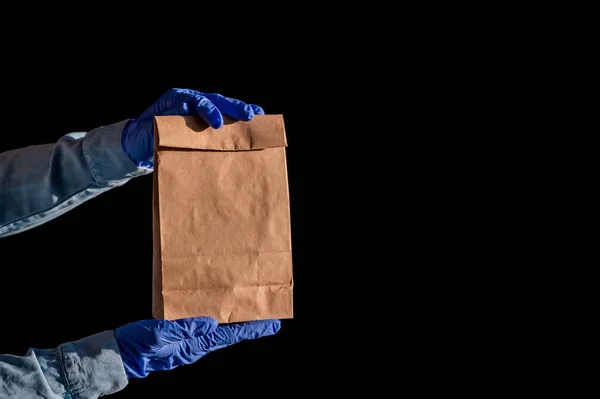 Weibliche Hände in einem blauen Handschuh halten eine braune Papiertüte vor schwarzem Hintergrund. Sichere Essenslieferung nach Hause. Ein Kurier im Jeanshemd reicht einem Kunden eine Bastelkartontasche. — Stockfoto