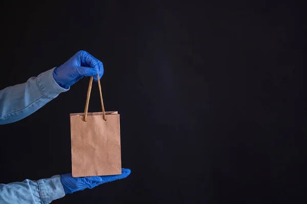 Weibliche Hände in blauen Handschuhen halten eine braune Papiertüte mit Henkeln auf schwarzem Hintergrund. Sichere Essenslieferung nach Hause. Ein Kurier im Jeanshemd reicht einem Kunden eine Bastelkartontasche. — Stockfoto