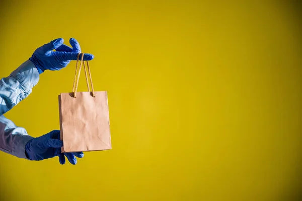 Female hands in gloves holds a small brown paper bag with handles on a yellow background. Safe food delivery to your home. A courier in a denim shirt holds out a craft cardboard bag to a customer. — Stock Photo, Image