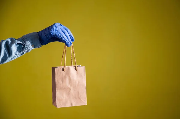 Fecho de mãos femininas em luvas e uma camisa de ganga. Um entregador está segurando um pequeno saco de papel junto às canetas em um fundo amarelo. Embalagem artesanal para lanche takeaway. Protecção antimicrobiana . — Fotografia de Stock