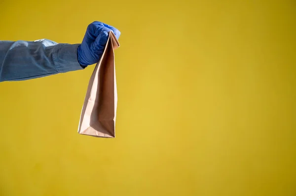 Closeup of female hands in gloves and a denim shirt. Delivery man passes an empty paper bag on a yellow background. Craft packaging for takeaway snack. Antimicrobial protection. — Stock Photo, Image