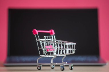 İnternetten alışveriş konsepti. Mini market arabası ve pembe arka planda dizüstü bilgisayar. Küçük araba ve kişisel bilgisayar.