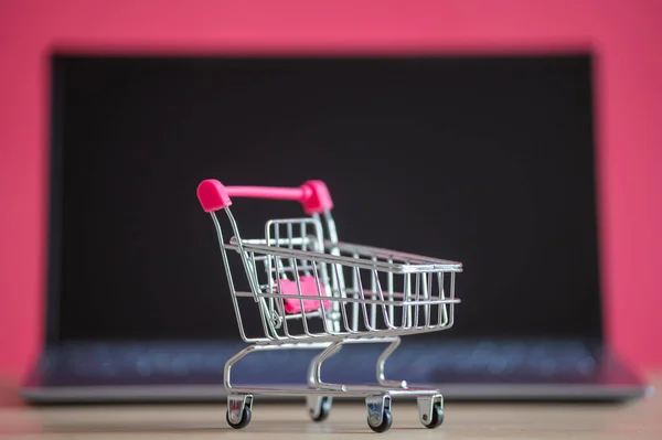 Conceito de compras online. Mini carrinho de supermercado e laptop em um fundo rosa. Carrinho pequeno e computador pessoal . — Fotografia de Stock